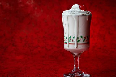 A DIY McDonald's egg nog milkshake, topped with whipped cream and a maraschino cherry in a glass holiday cup, on a red background.