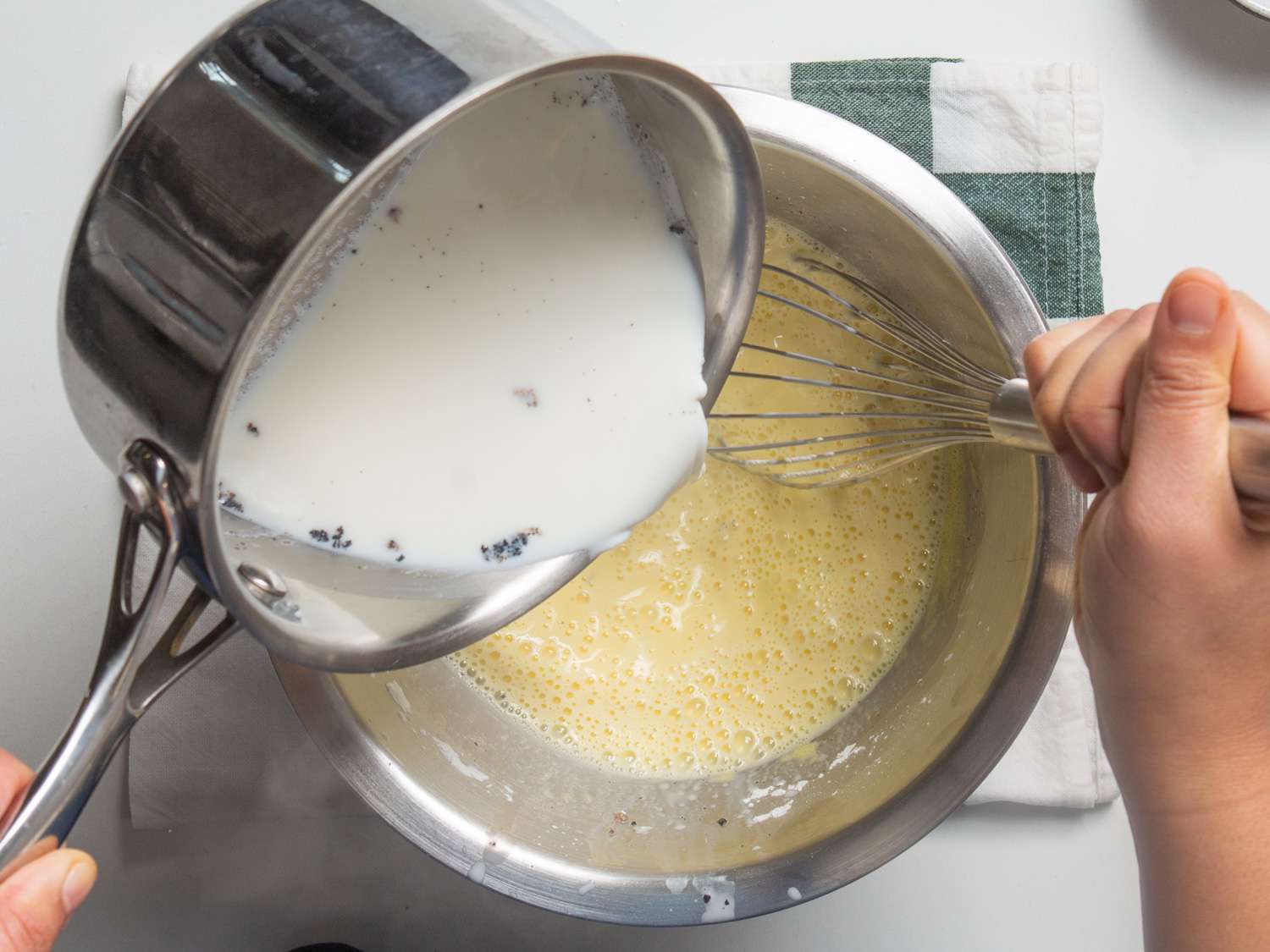 One hand pouring milk from saucepan into bowl with yellow custard and hand holding a whisk.