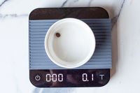A coffee bean in a small white bowl placed on Acaia Pearl showing 0.1 grams