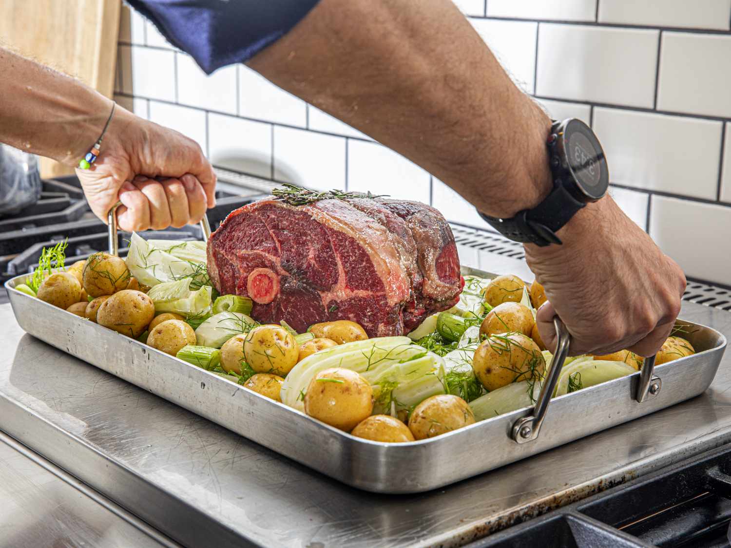 two handles grabbing onto a roasting pan's handles (the roasting pan has meat, potatoes, and fennel in it)