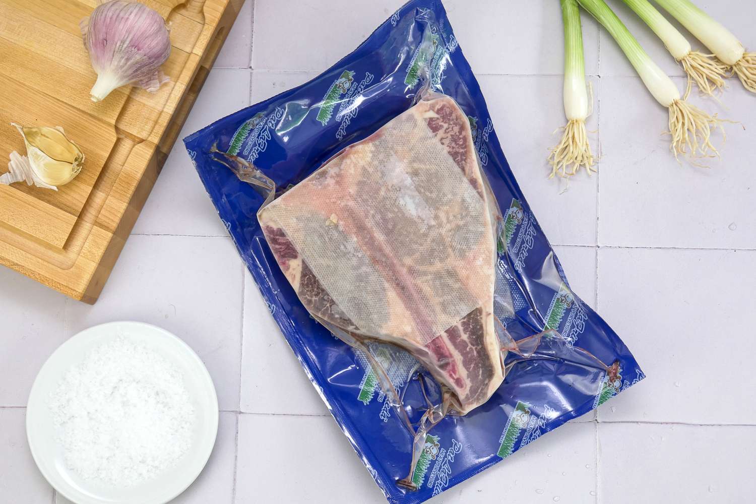 A packaged piece of meat on a countertop with garlic scallions and a bowl of salt