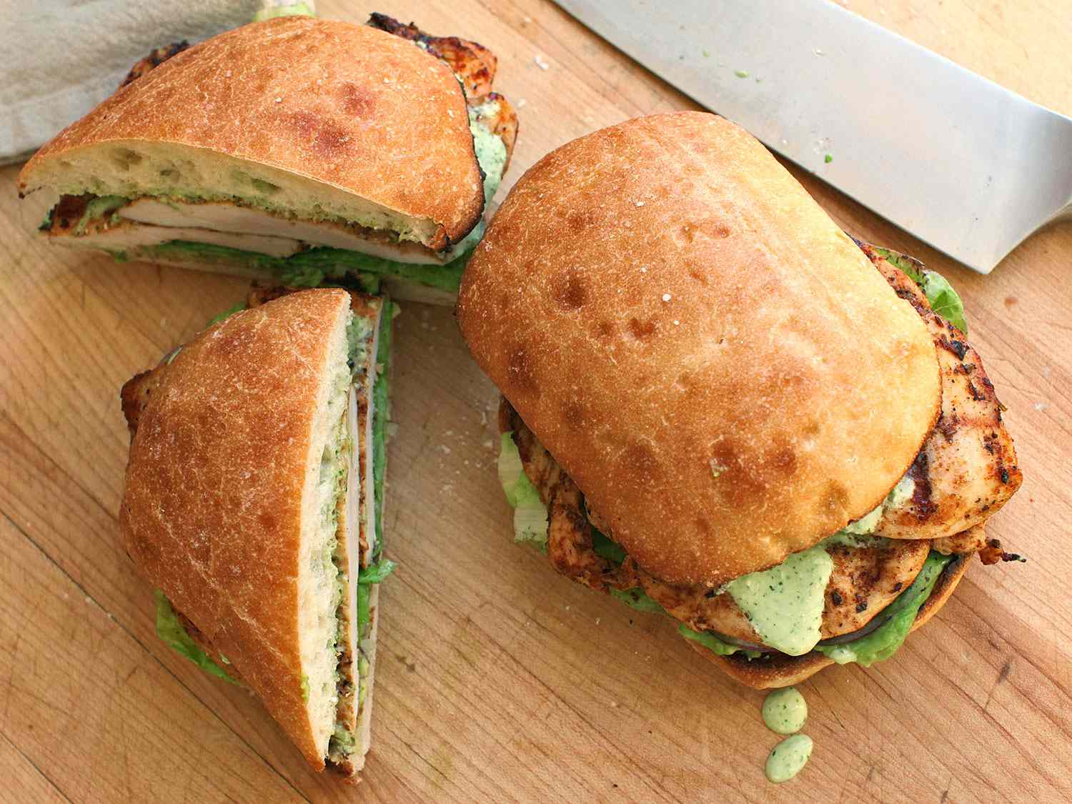 A pair of Peruvian chicken sandwiches, one cut on the diagonal, the other dripping creamy jalapeño sauce onto a wooden cutting board.