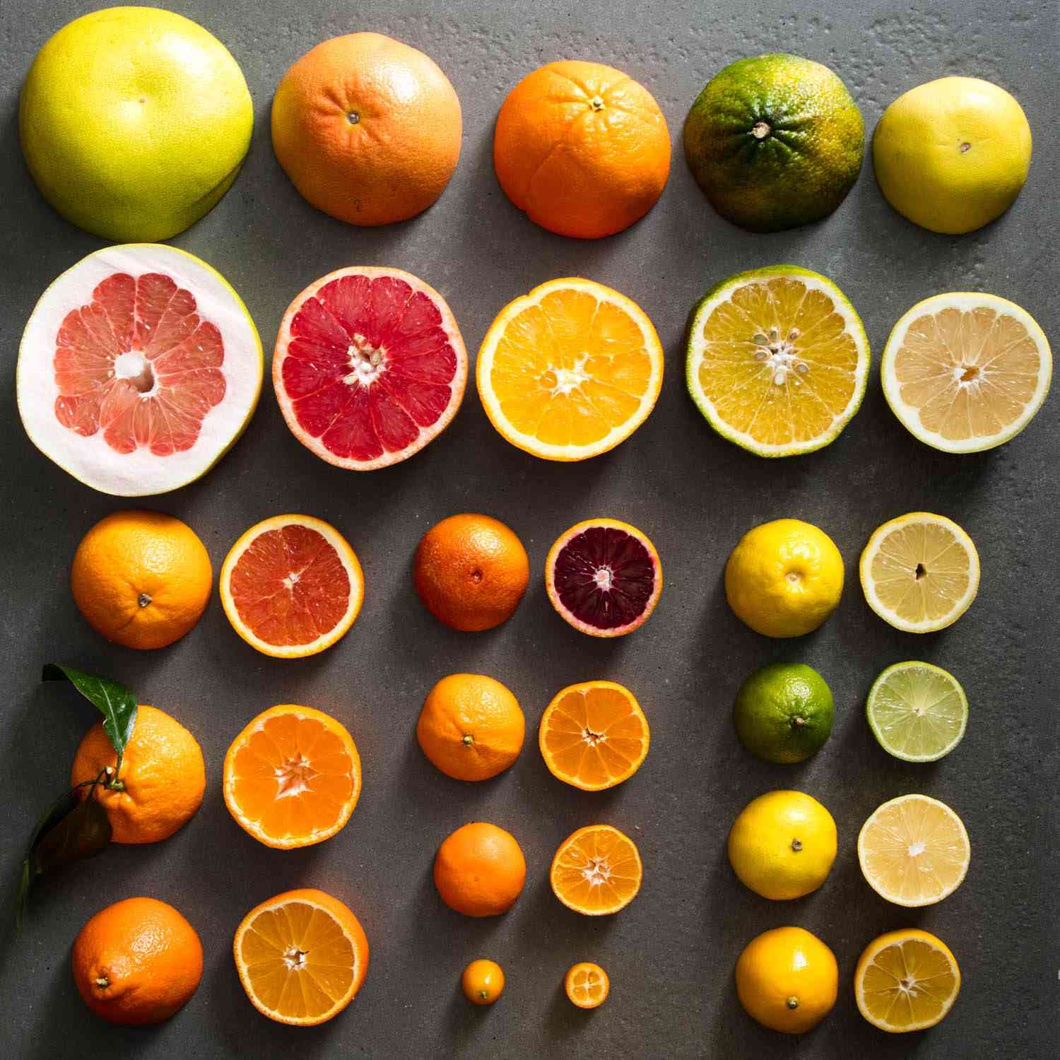 A line up of whole and halved citrus, including pink, thick-skinned pomelos, ruby grapefruits, oranges, lemons, limes, kumquats and more.
