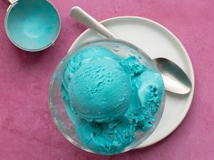 Blue moon ice cream in a glass bowl, set on a white ceramic plate which also holds a metal spoon. The background is pink and in the top left of the image is the ice cream scoop which had been used to scoop the ice cream.