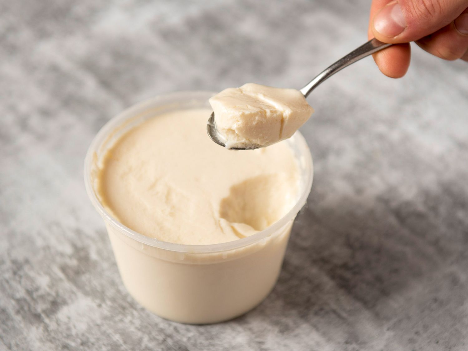 Hand holding spoonful of fresh tofu lifted from plastic container