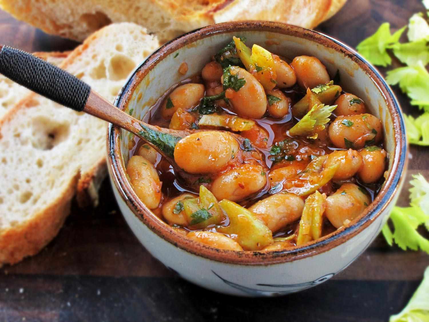 A bowl of giant Spanish beans next to a baguette and sliced celery leaves.