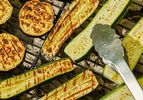 Slices of zucchini and eggplant grilling on a barbecue with metal tongs