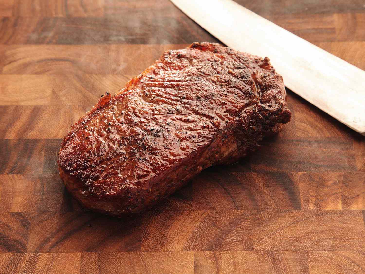Cooked steak resting on a cutting board with a knife off to the side. 