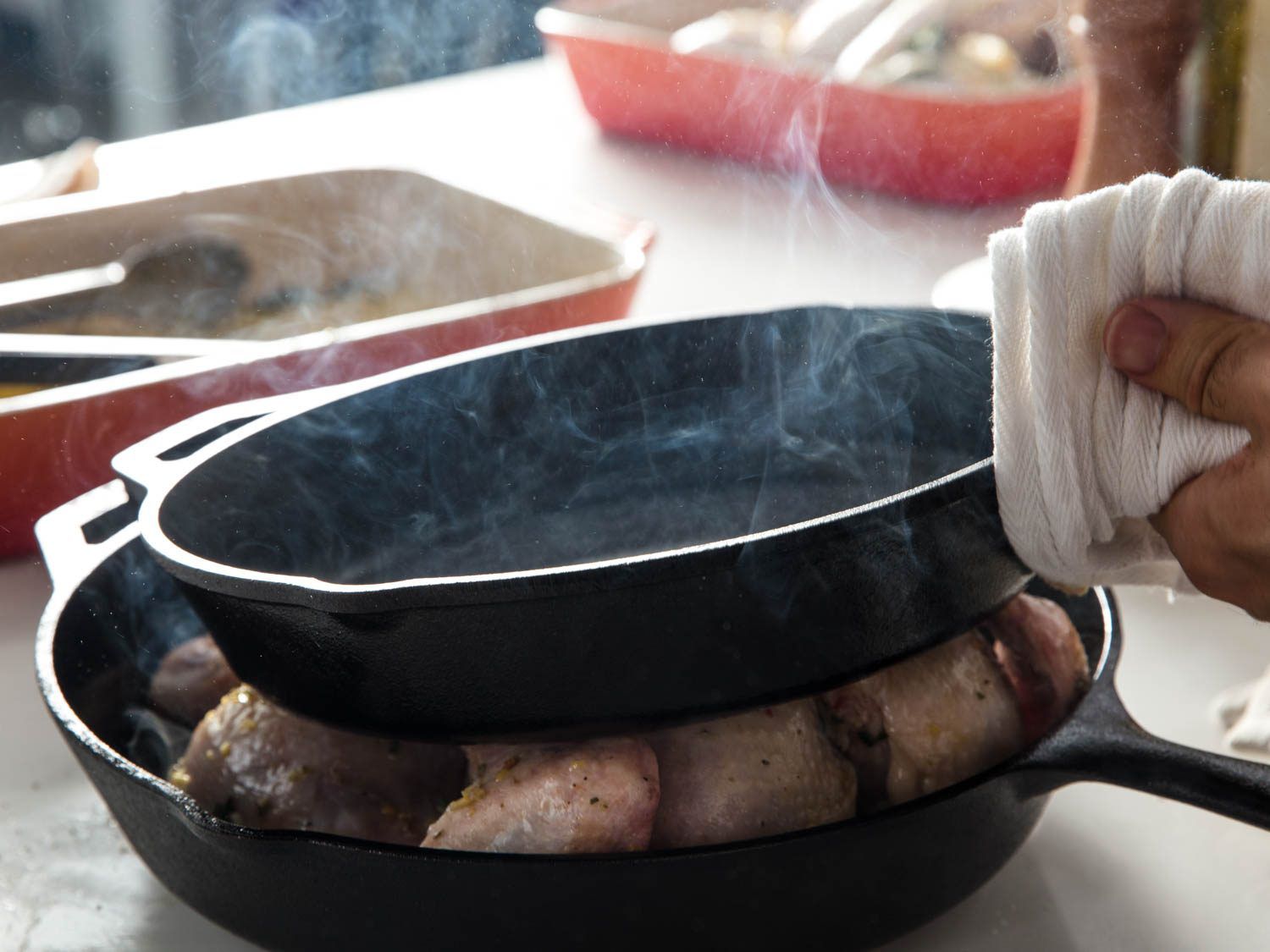 A smoking-hot cast iron skillet being lifted off a spatchcocked chicken in another cast iron skillet.