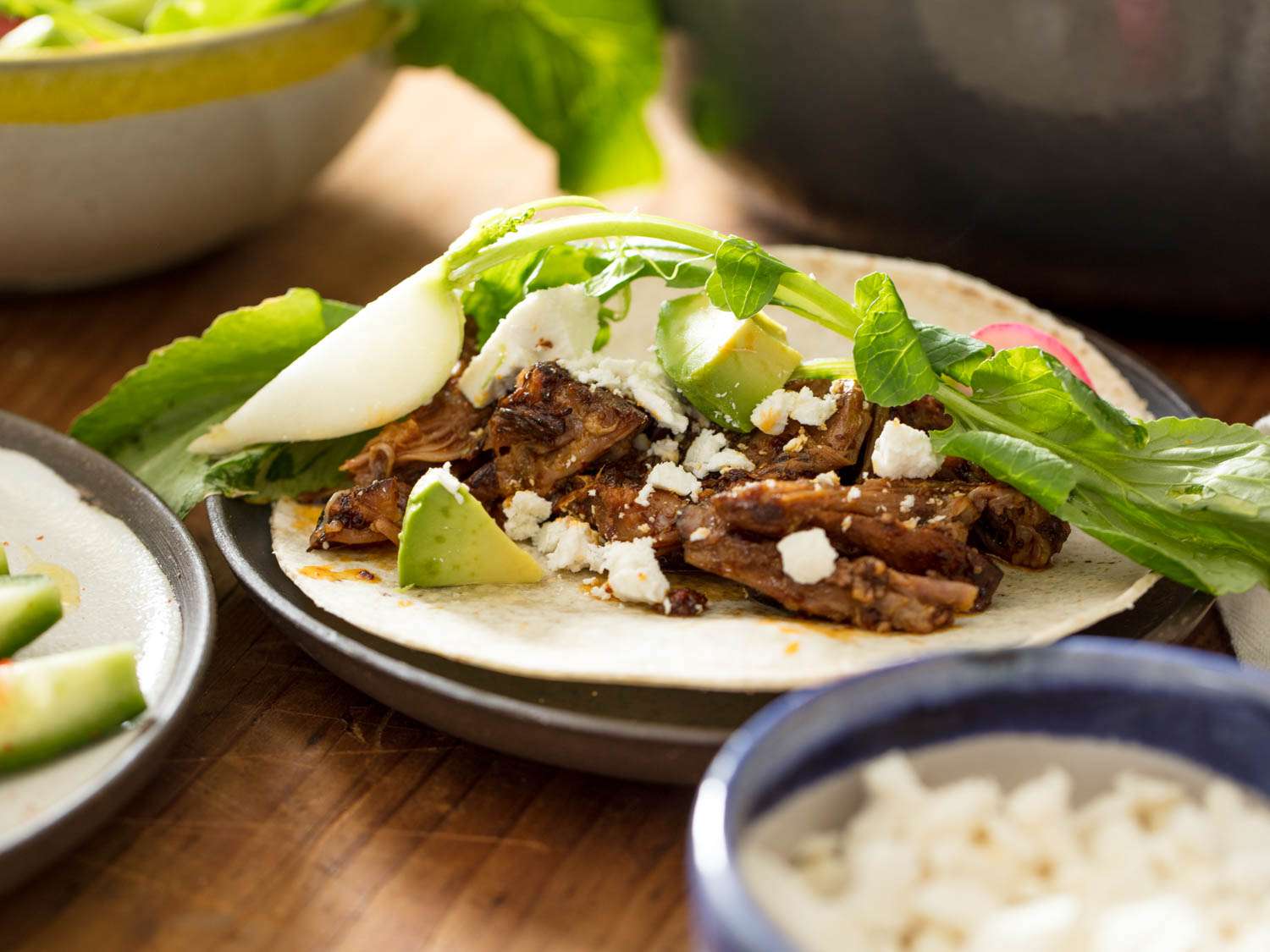 Braised Lamb Shoulder With Dried Chiles and Dates, served as a taco with cotija, avocado, radish slices, and a quartered hakurei turnip and its green top.