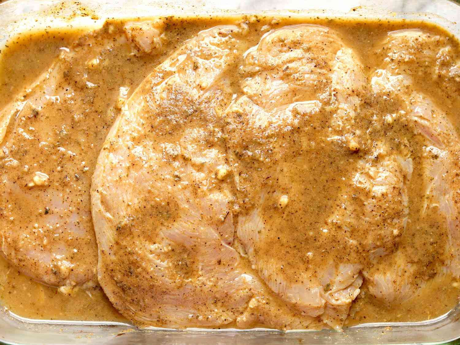 Raw skinless chicken breasts in a container, coated with marinade. 