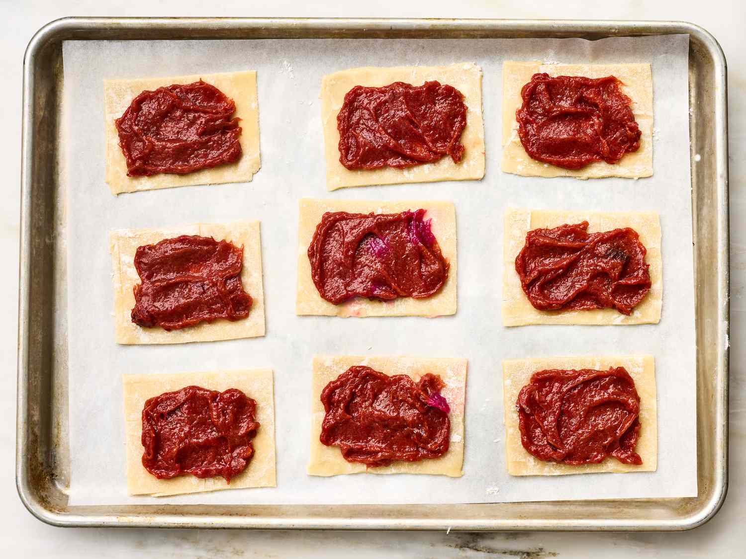 Nine rectangles of dough topped with red strawberry-rhubarb filling on a baking tray lined with parchment paper