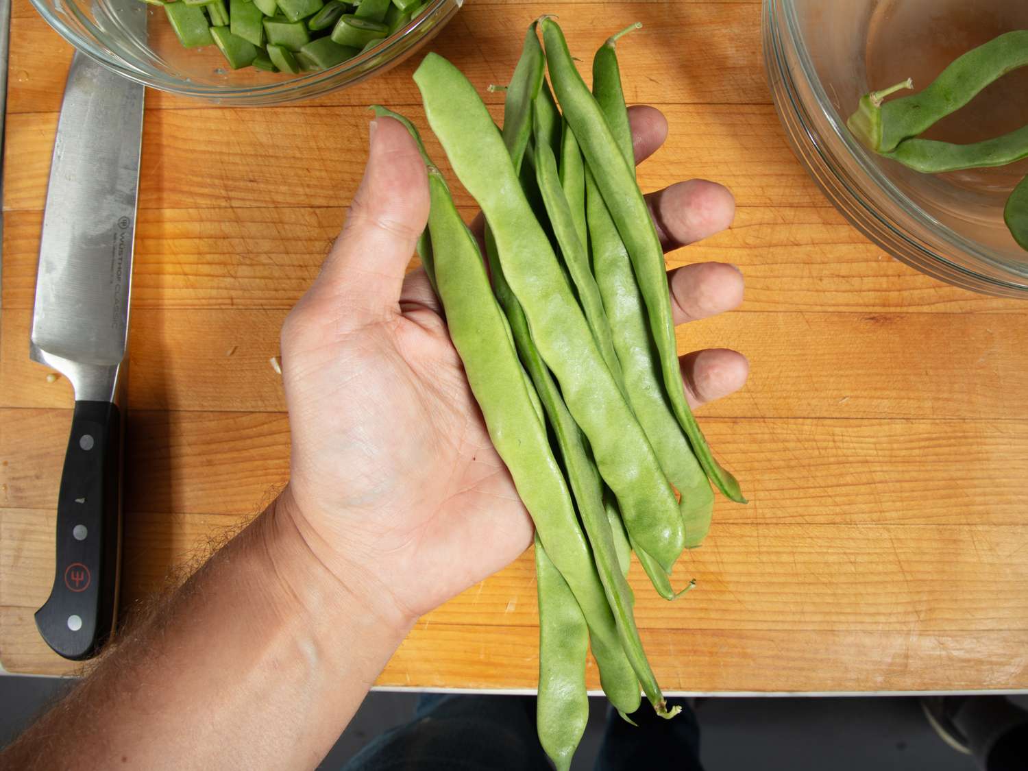 A hand holding several green beans over a wooden surface with a knife and bowls nearby