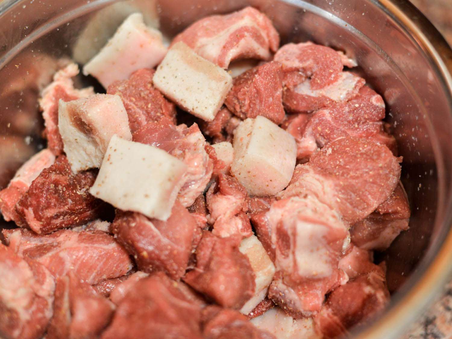Chunks of lamb, pork, and fatback are seasoned and tossed in a mixing bowl.