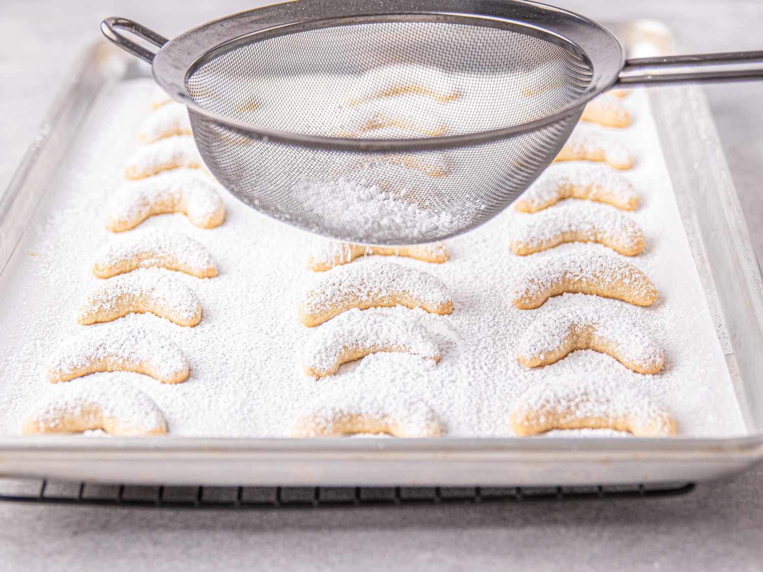 Powdered sugar being sifted over a baking tray of crescentshaped cookies