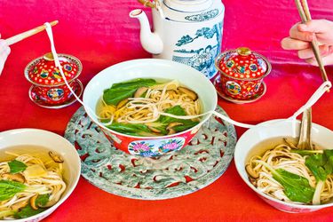 A large bowl of longevity noodles are served on a red table, alongside a pot of tea. Diners serve themselves from the bowl, transferring the long noodles with chopsticks.