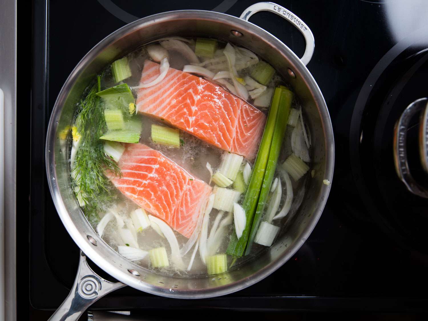 An overhead shot of a saucepan containing two pieces of raw salmon fillet, sliced fennel, celery, leeks, a bay leaf, and several sprigs of dill.