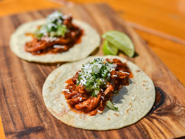 Two chicken tinga tacos on a woode n serving board, garnished with tomatillo salsa, cilantro, onion, and grated cotija. Lime wedges are served alongside.