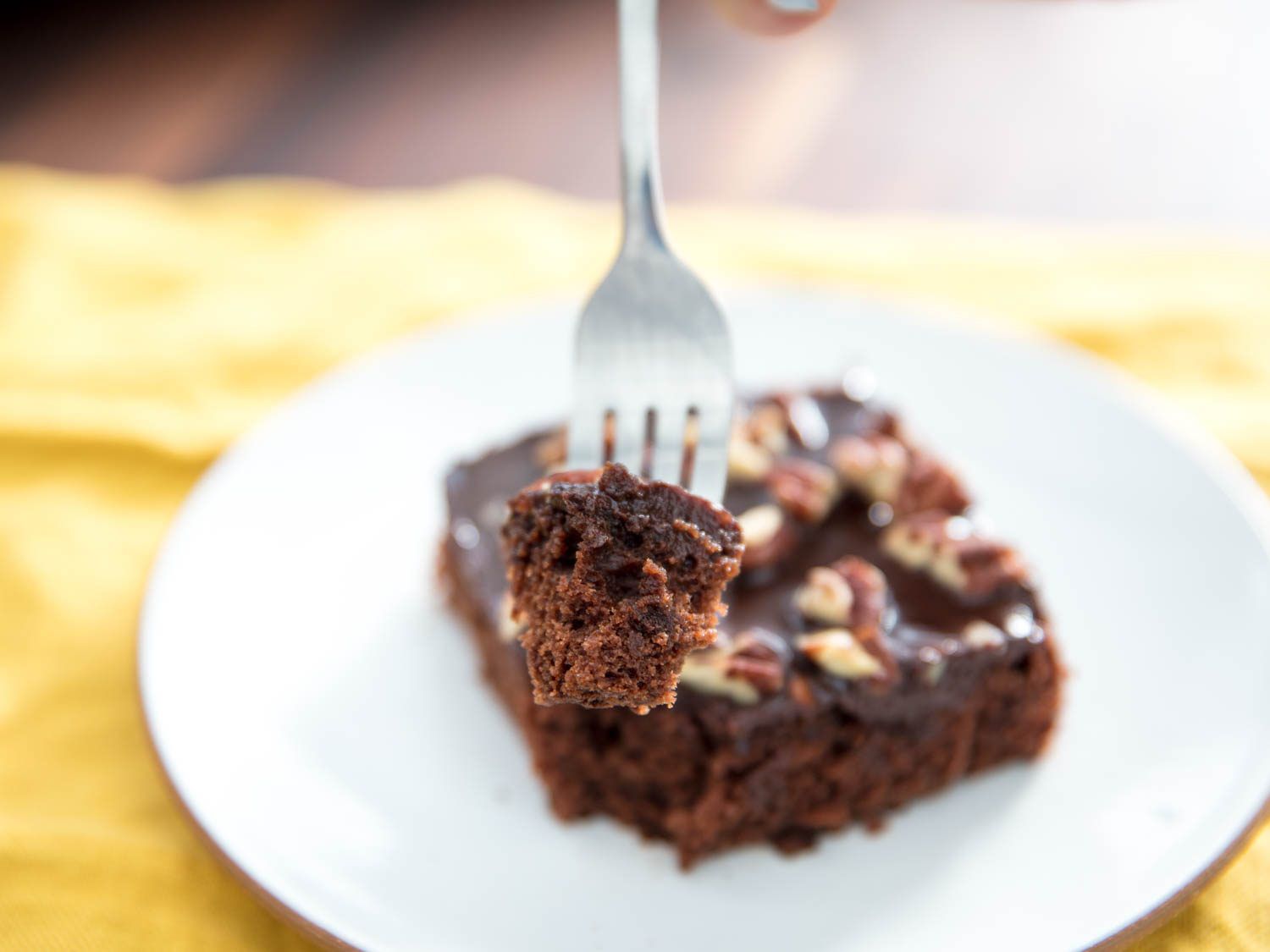 A bite of Texas sheet cake speared on a fork, with a square of cake on a white plate in the background.