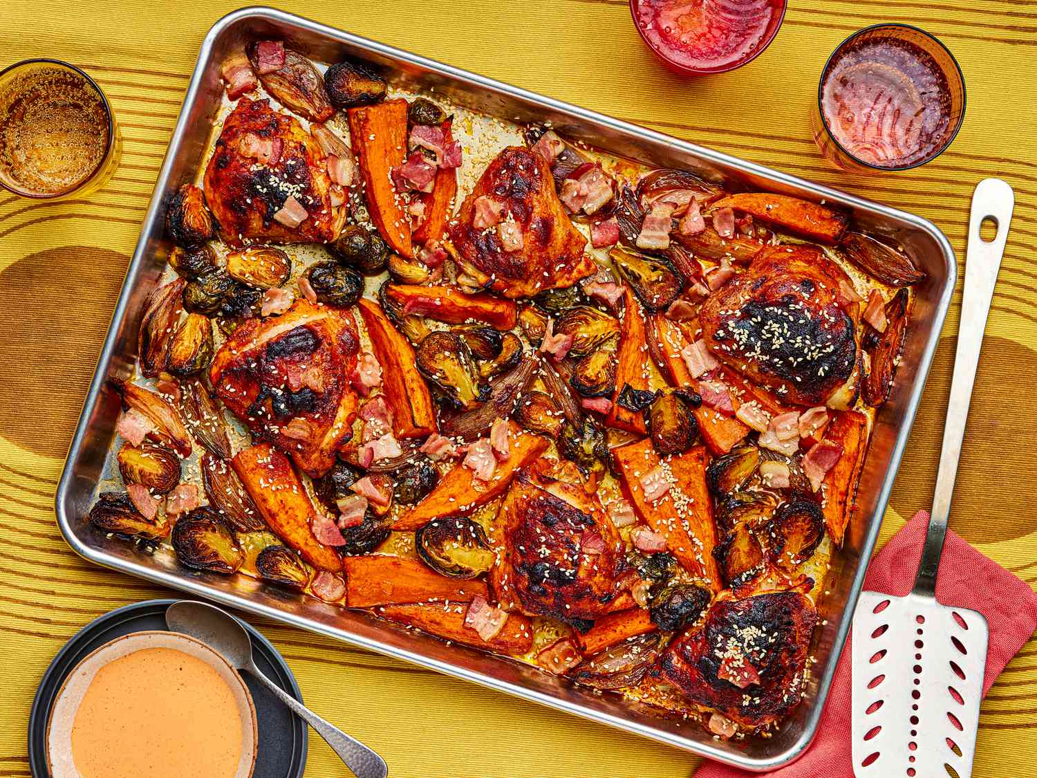 Sheet pan of roasted chicken and vegetables on a yellow and brown surface, with red cups and a serving spatula
