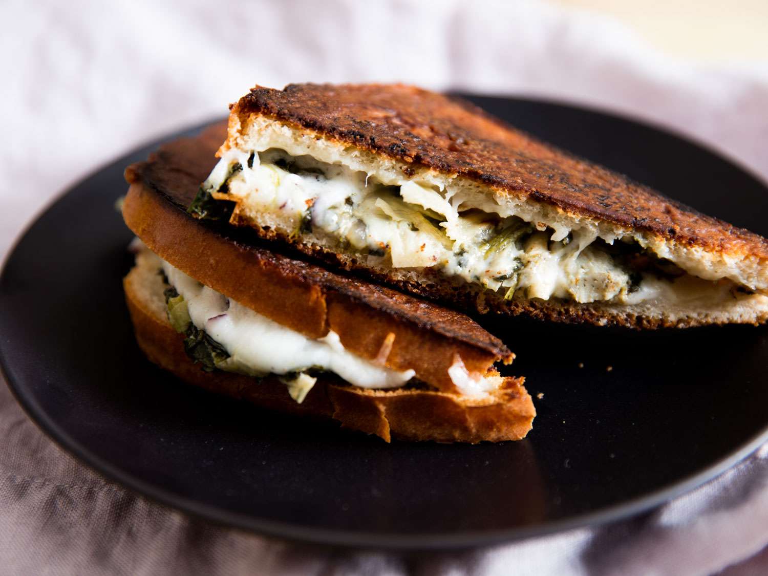 A crisp grilled cheese sandwich stuffed with spinach and artichokes, cut in half and stacked on a black plate.