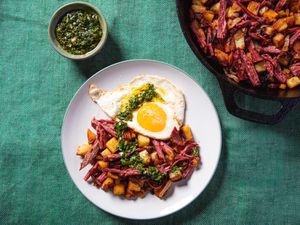 Plated Corned Beef Hash topped with fried egg and chimichurri, flanked by skillet of corned beef hash and dish of chimichurri 