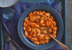 Glazed carrots in a blue bowl on blue rustic surface. A spoon is in the bowl, with a few carrot slices in it, and there is cups of sparkling water, and plates on the side.