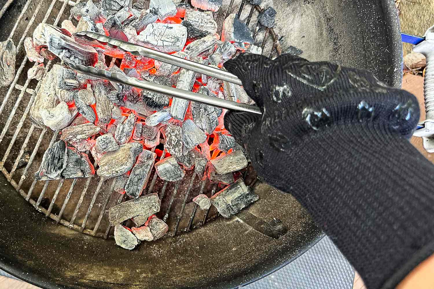 A hand in a heatresistant glove holding metal cooking tongs over smoldering charcoal in a grill