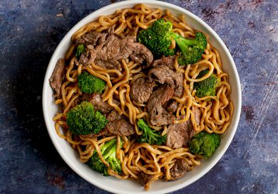 A white ceramic bowl holding lo mein with beef and broccoli on a purple colored stone surface.