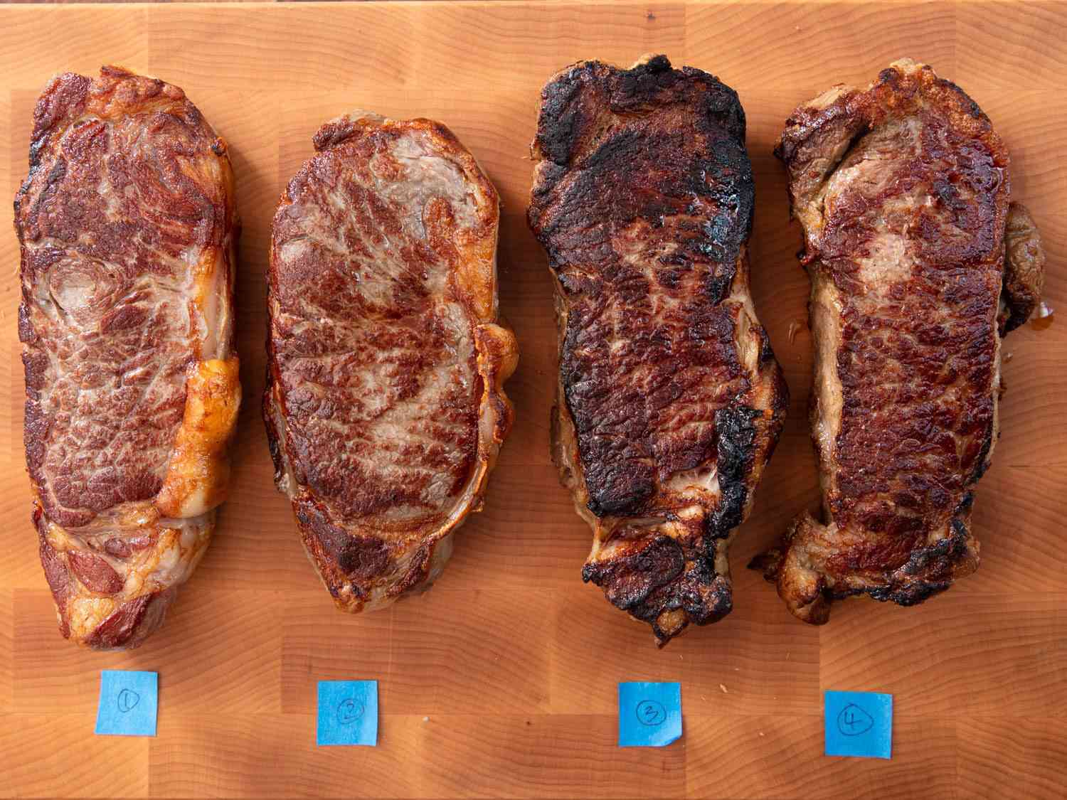 Overhead view of four strip steaks cooked sous vide then seared, identified by numbered tape on a cutting board. Steak number three, the one that had been coated in Skippy, is visibly darker, and a bit burnt.