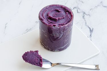 A glass jar of ube halaya placed on a white marble surface. A morsel of the stiff jam is perched on a spoon in front of the jar.