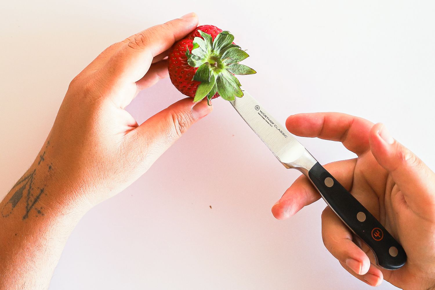 Hands holding a strawberry and a Wüsthof knife with the knife positioned against the stem of the strawberry for cutting