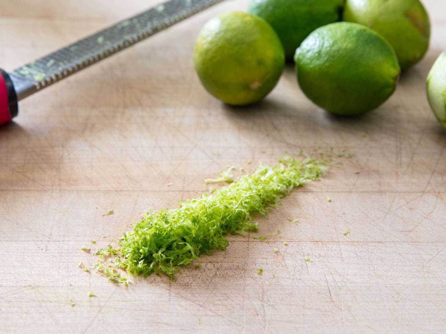 A pile of lime zest sits on a cutting board next to several whole limes and a microplane zester.