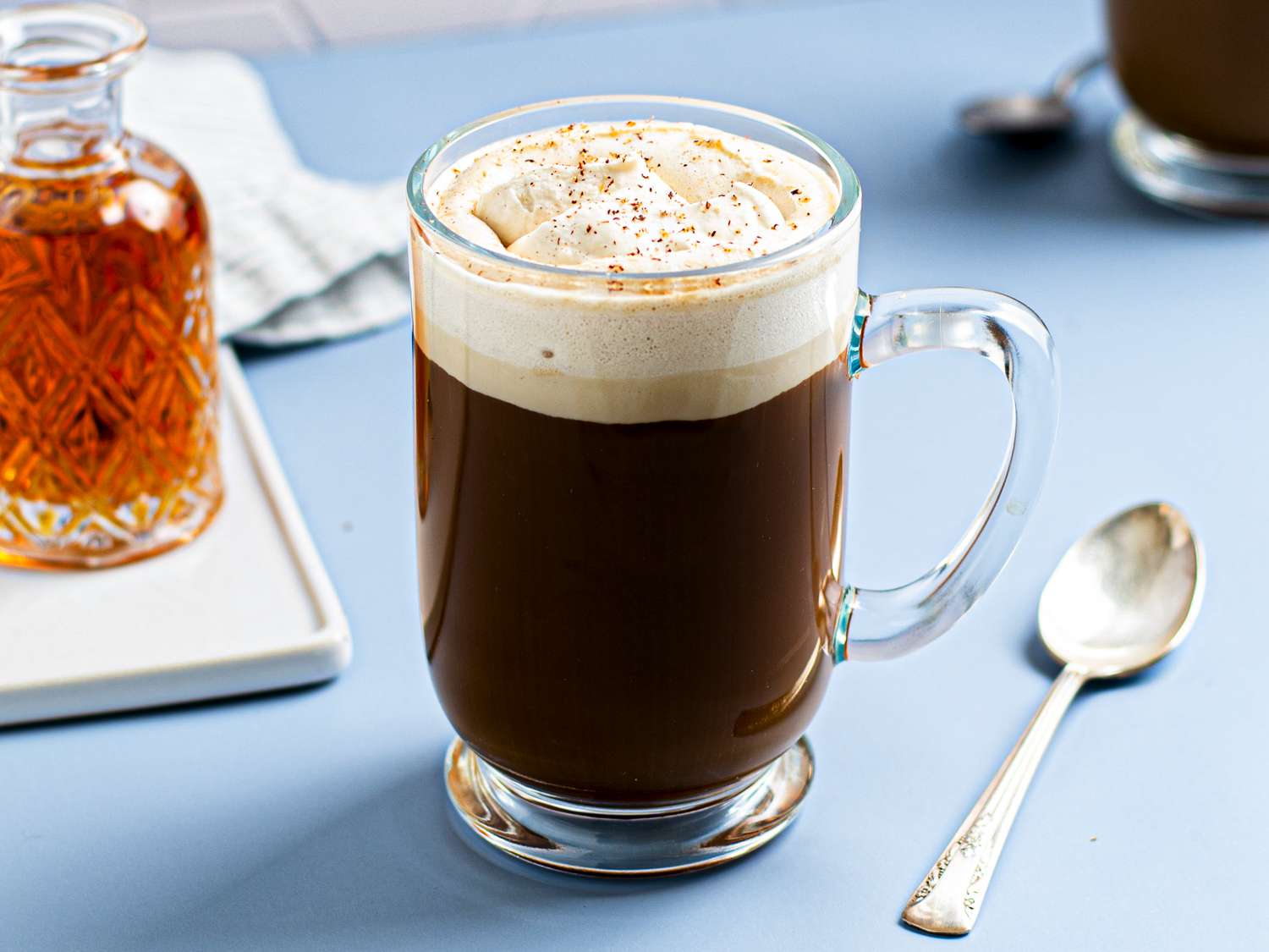 Spiced rum coffee with butterscotch whipped cream in a glass mug. There is a silver spoon to the right of the mug, and a small glass vessel of rum on the lefthand side of the image.