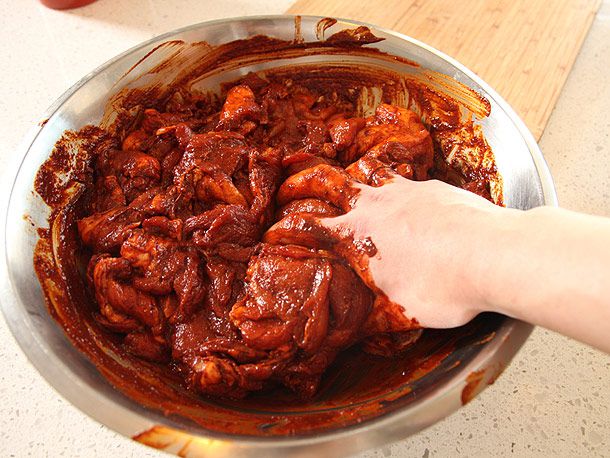 A hand mixing red chile marinade into raw pork in a large metal bowl