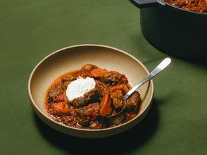 Bowl of beef stew with vegetables and sour cream served with a spoon pot of stew in the background