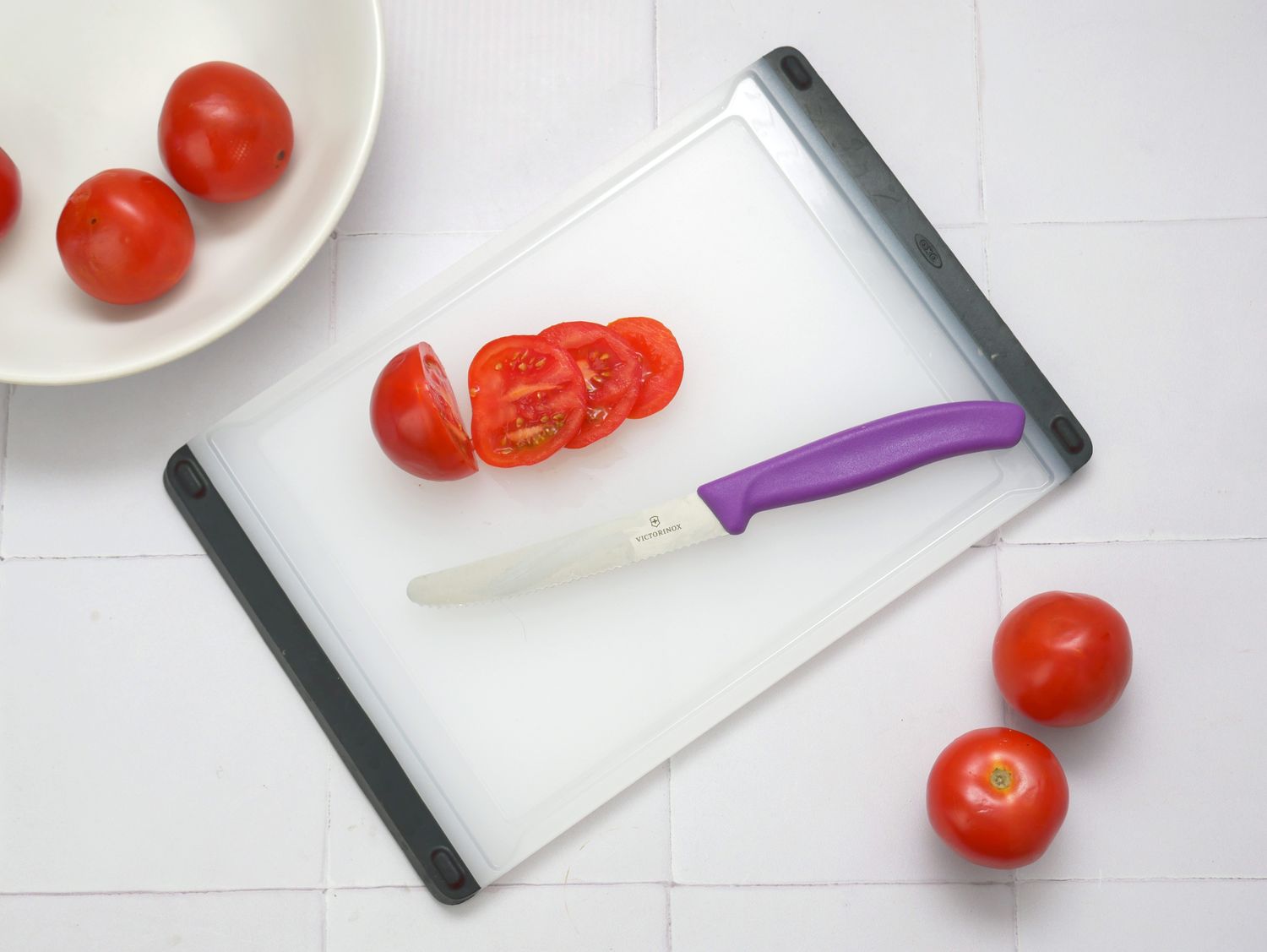 A serrated paring knife with a purple handle slicing tomatoes on a cutting board with additional tomatoes nearby