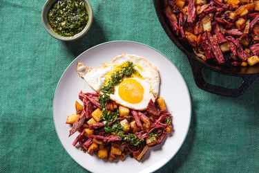 Plated Corned Beef Hash topped with fried egg and chimichurri, flanked by skillet of corned beef hash and dish of chimichurri 