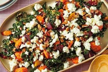 A salad made with kale roasted root vegetables and crumbled cheese topped with nuts on a rectangular serving dish