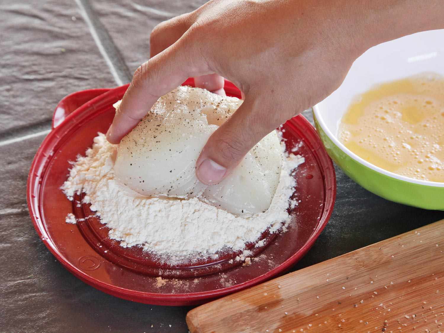Author pressing a seasoned fish fillet portion into a dish of flour.