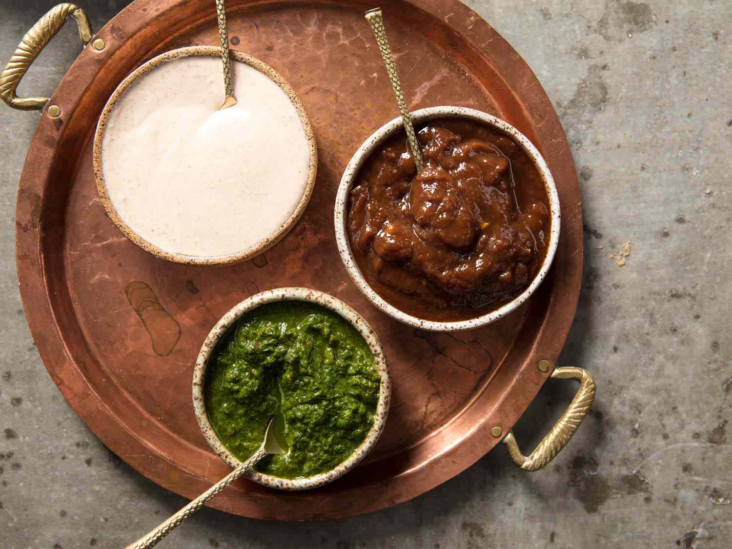 A copper tray holding three small bowls. One bowl holds spiced yogurt, one holds tamarind chutney, and one holds mint chutney.