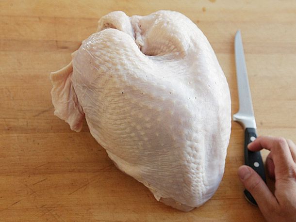 A whole bone-in turkey breast on a wooden cutting board with a hand holding a boning knife on the side.