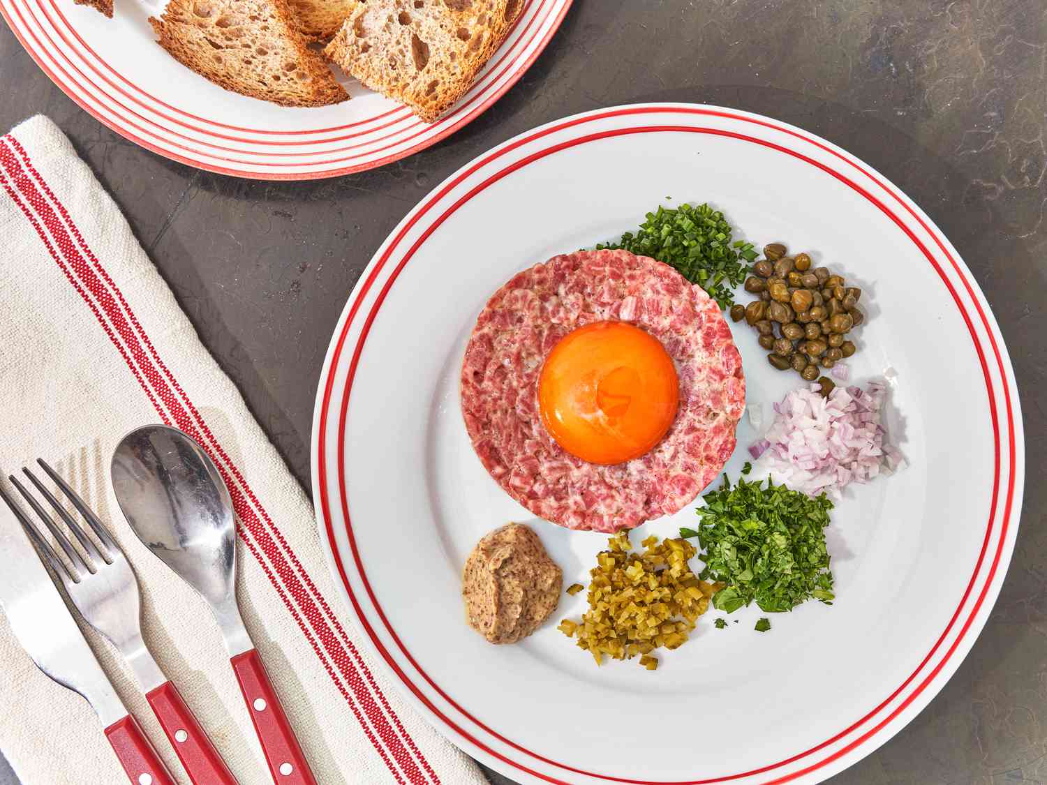 Beef tar tar with capers, onions and accoutrements on a dish with utensils, napkin, and plate of toasted bread around it