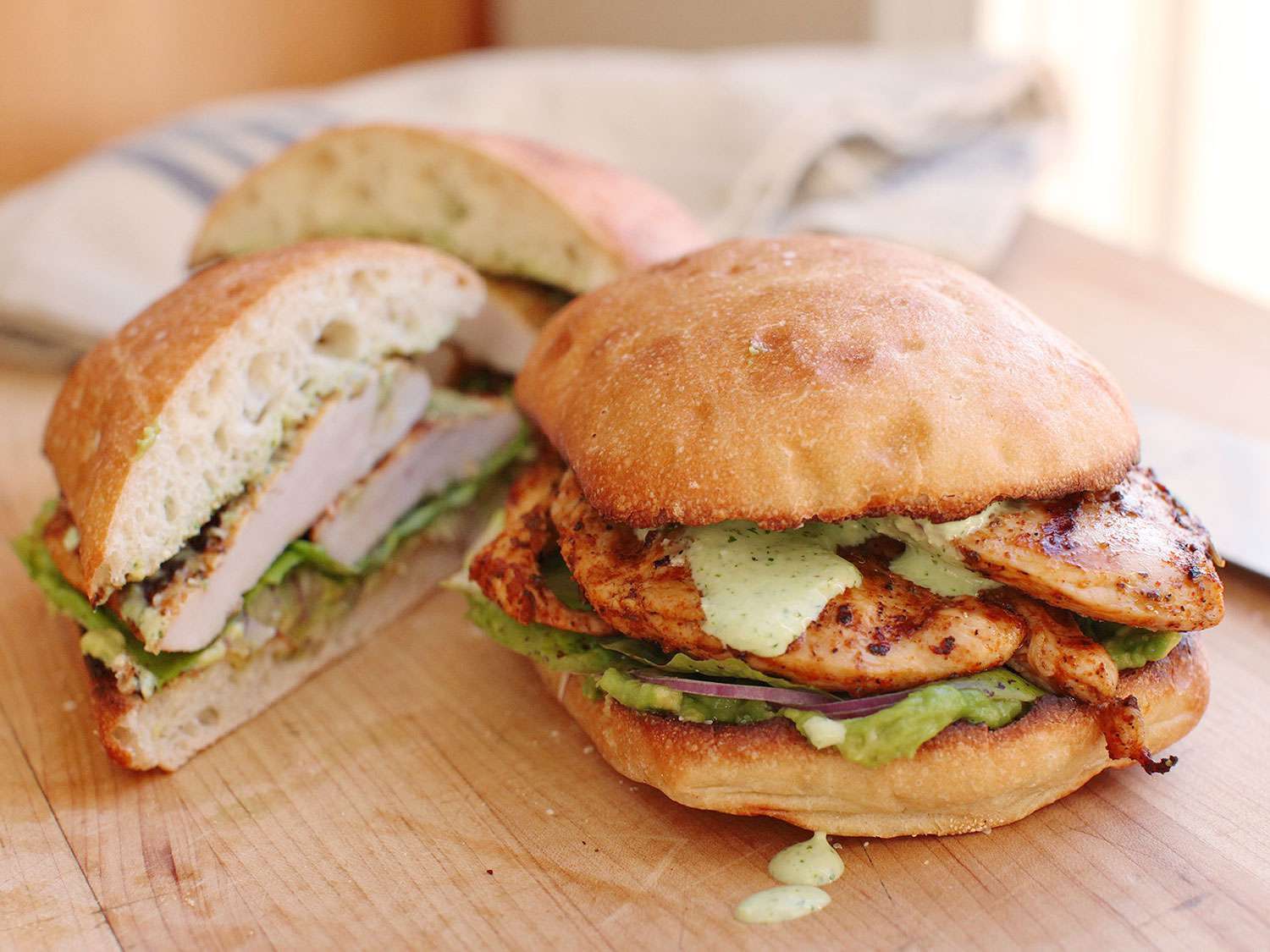 A Peruvian grilled chicken sandwich topped with avocado, red onion, and green sauce, with two sandwich halves nearby.