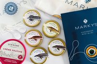 A collection of products from Marky's, including beluga, kaluga, siberian sturgeon, osetra, and white sturgeon caviars, blinis, a tote bag, and crème fraîche