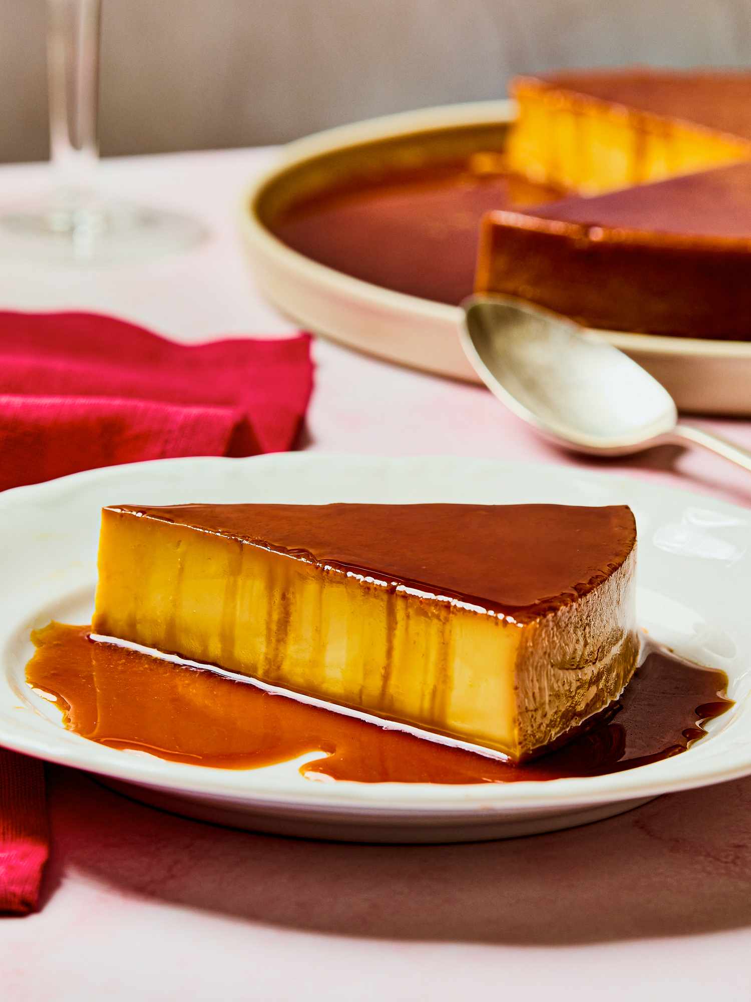A slice of carameltopped flan on a plate with the rest of the dessert in the background