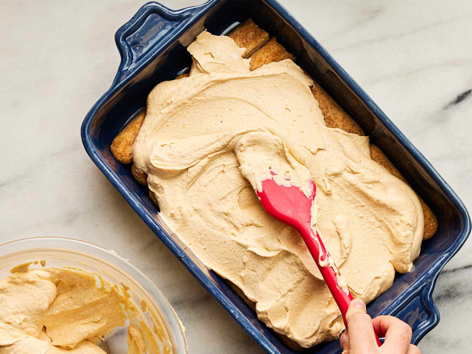 spreading mascapone onto lady fingers in a baking dish, with bowl of mascapone on the side 