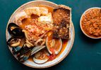 A serving of cioppino in a bowl, overflowing with plump shrimp, mussels, clams, calamari, fish, and more. There's a piece of deeply toasted sourdough on the side, and a small bowl holding a roasted red pepper condiment.