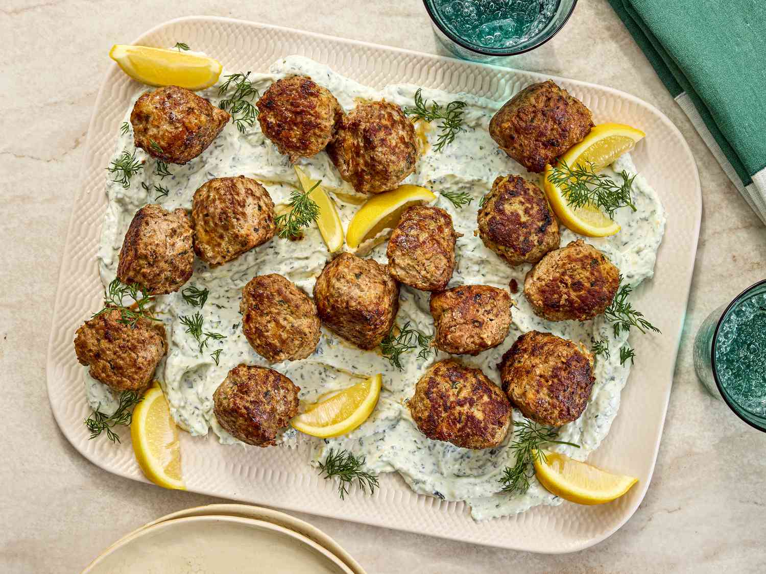 meatballs on top of yogurt with fresh herbs and lemon wedges on a platter. Side of sparkling water and green napkin on the side 