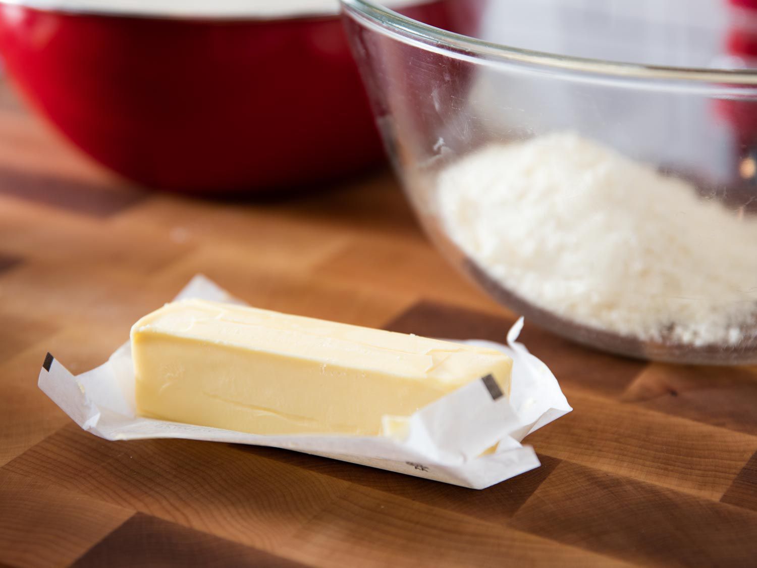 An unwrapped stick of butter on a wooden cutting board. A bowl of grated Parmigiano-Reggiano cheese is on the background. 